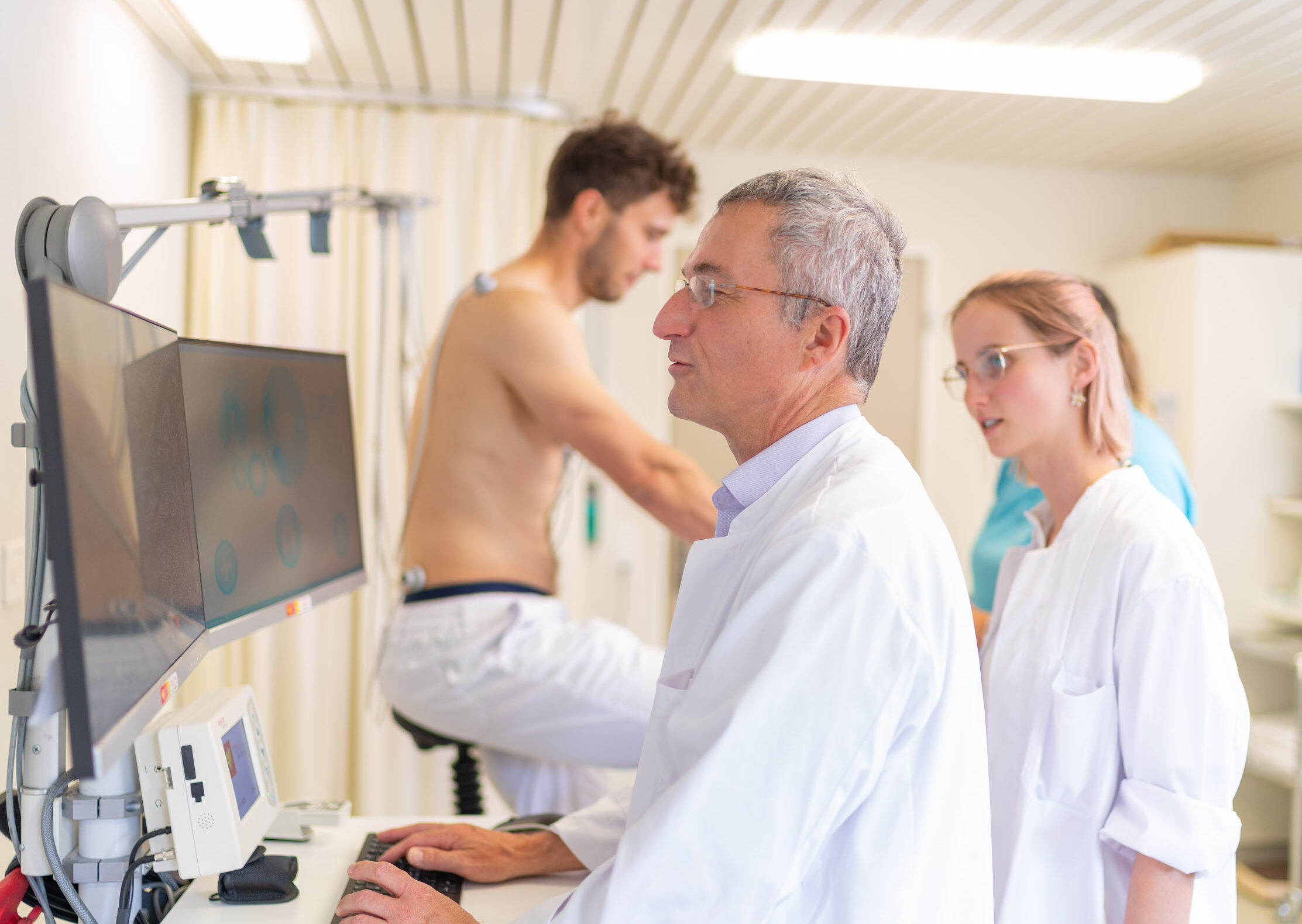 Ärzte am Bildschirm, Patient im Hintergrund auf Ergometer